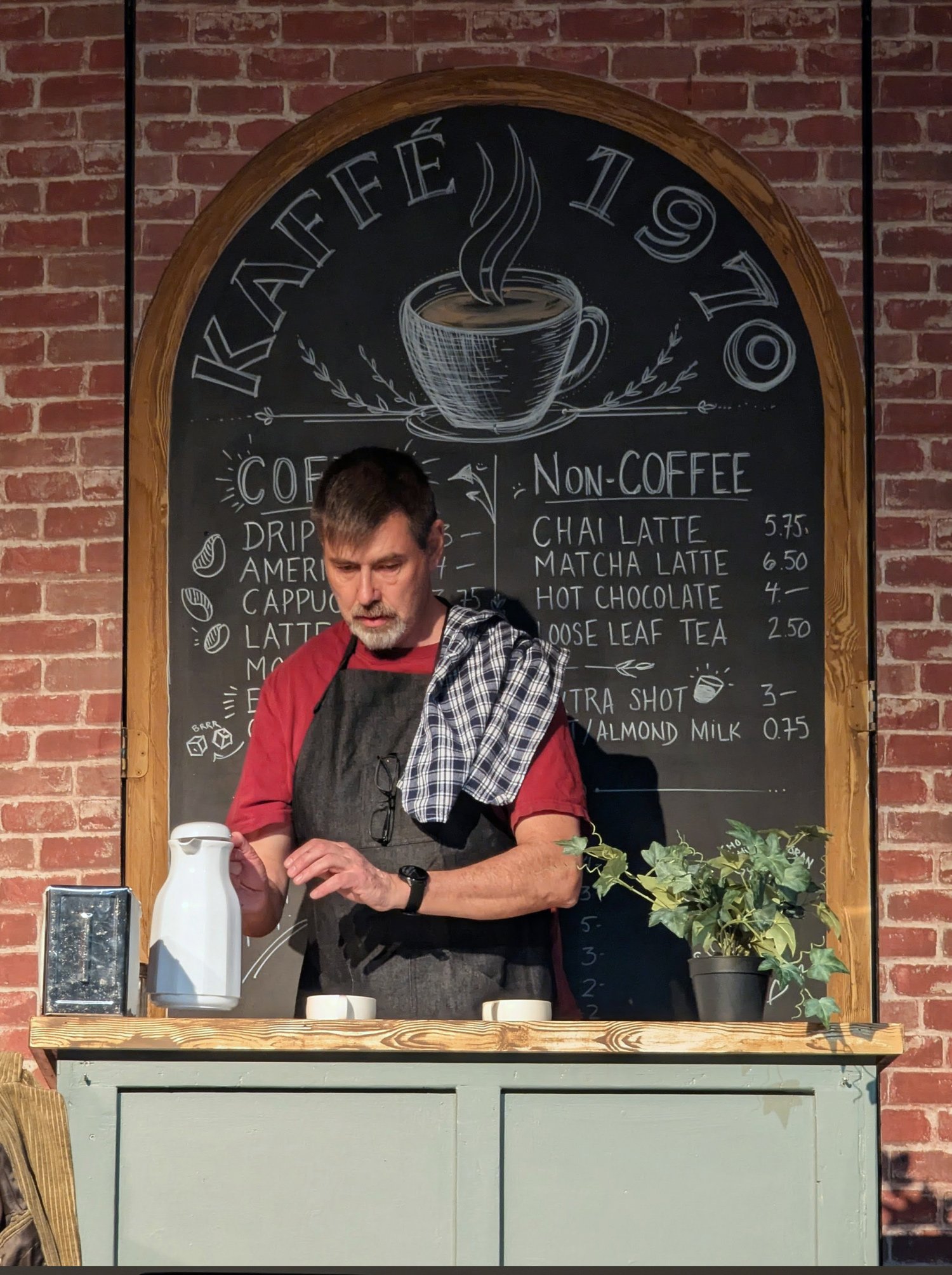 Barista behind a caf&eacute; counter set with a hand-chalked Kaffe 1970 menu board on a brick wall